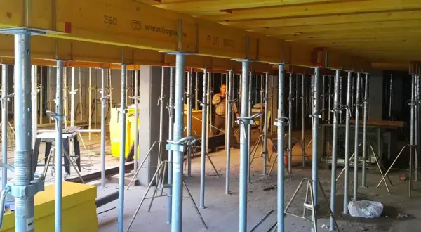Metalowe stemple budowlane podtrzymują żółte belki stropowe. W tle widać pracownika i inne elementy konstrukcyjne.