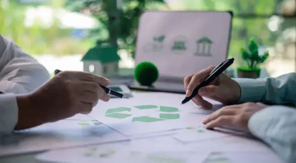 Manos revisando documentos con símbolo de reciclaje y un portátil con iconos de sostenibilidad, preparándose para una certificación verde.