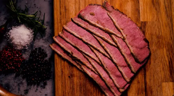 Cienkie plastry różowego pastrami na desce, obok przyprawy. Zastanawiasz się, pastrami co to? To aromatyczna, peklowana wołowina.