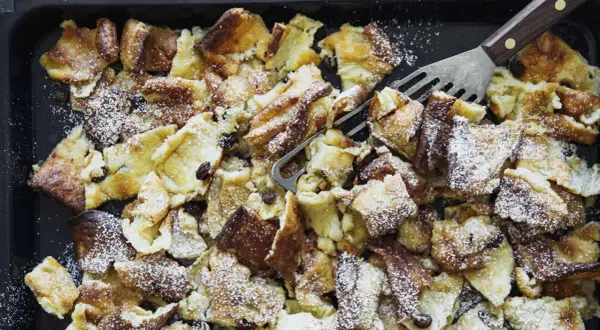 Kaiserschmarrn aus dem Backofen auf einem Blech, mit Puderzucker bestäubt und Rosinen. Ein Gabel-Schaber liegt bereit.
