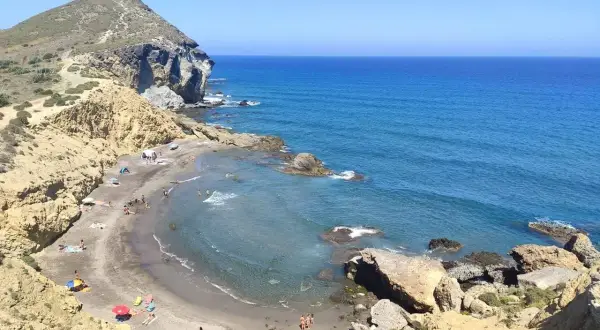 Vista aérea de **playas nudistas Cabo de Gata**. Arena oscura, aguas turquesas y formaciones rocosas. Gente disfrutando del sol y el mar.