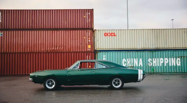 Klasyczny zielony **hatchback** Dodge Charger z białym pasem na tyle, zaparkowany przy stosach kontenerów w porcie.