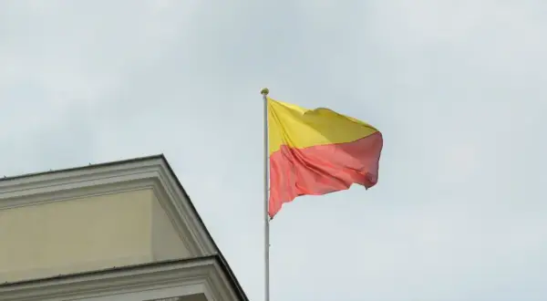 Flaga Warszawy powiewa na maszcie obok zabytkowego budynku.