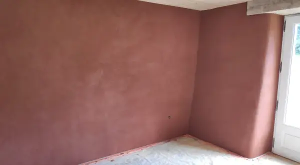 Un mur d'angle avec un enduit chaux sable intérieur d'une belle teinte ocre, sous un plafond en bois clair. Le sol est protégé par une bâche.