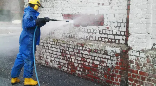 Personne en combinaison bleue et bottes jaunes utilise un nettoyeur haute pression pour enlever peinture sur brique.