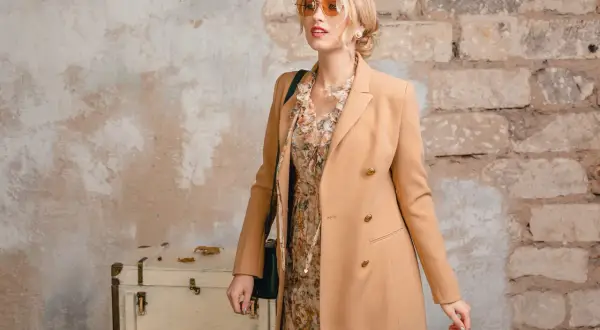 Elegancka kobieta w płaszczu w kolorze camel, sukience w kwiaty i okularach przeciwsłonecznych pozuje przy starej walizce.