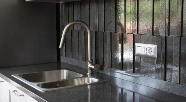 Top cucina in pietra lavica: pro e contro. Un lavello doppio in acciaio inox e un rubinetto moderno su un piano scuro e un rivestimento a listelli verticali.