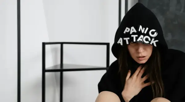 Kobieta w czarnym kapturze z napisem "PANIC ATTACK" na okularach, trzymająca się za szyję, przeżywa uczucie umierania.