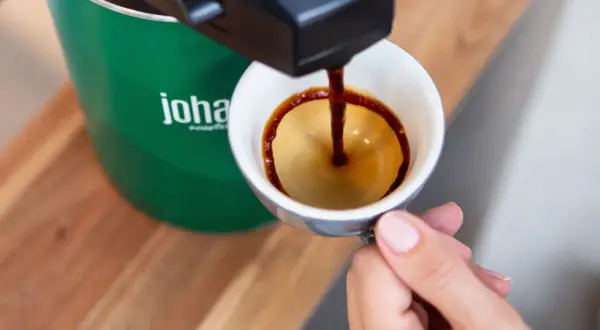 En hand håller en kopp medan kaffe rinner ner från en maskin. Frågan om hur många matskedar kaffe per kopp är relevant för att få den perfekta smaken.