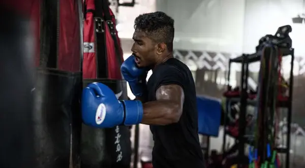 Boxer in blue gloves throwing a powerful cross punch at a heavy bag, sweat glistening on his arms.