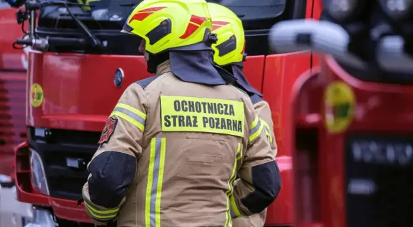 Strażacy w żółtych hełmach i beżowych kurtkach z napisem "Ochotnicza Straż Pożarna" stoją obok czerwonego wozu strażackiego.