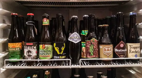 Scaffale pieno di bottiglie di birra, alcune con etichette colorate come "Zinnebir" e "Saison Dupont". Una birra aperta in frigo, pronta per essere gustata.