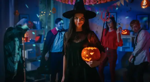 Kiedy obchodzi się Halloween, młoda kobieta w stroju czarownicy trzyma świecącą dynię na imprezie z przyjaciółmi w kostiumach.