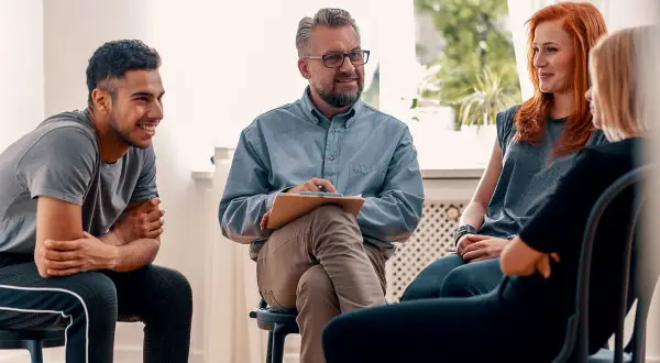 Grupa ludzi na sesji terapeutycznej. Mężczyzna z notatkami prowadzi rozmowę, a uczestnicy słuchają. Może uczą się języka francuskiego?