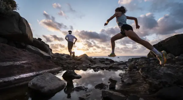 Deux coureurs s'entraînent sur un sentier rocailleux à Tenerife au coucher du soleil, l'océan en arrière-plan.