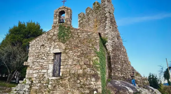 Antigua ermita de piedra con campanario y cruz, rodeada de vegetación. Las **torres de oeste** se alzan imponentes contra el cielo azul.