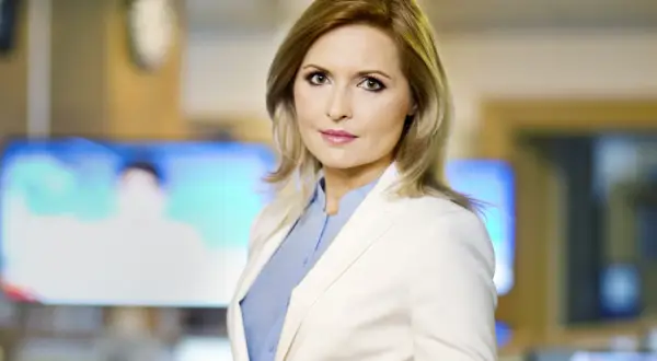 Agnieszka Gozdyra w białej marynarce i niebieskiej koszuli, w tle ekrany telewizyjne. Czy Agnieszka Gozdyra ma dzieci? Tego nie widać na zdjęciu.