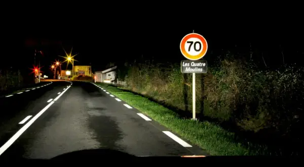 Route de nuit avec marquage au sol blanc lumineux. Panneau "Les Quatre Moulins" et limite de vitesse à 70.