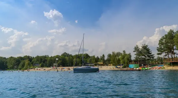 Żaglówka cumuje przy piaszczystej plaży nad jeziorem Głębocko Opolskie. Ludzie odpoczywają i kąpią się w słoneczny dzień.