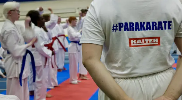 En grupp utövare i vita dräkter tränar karate. En person bär en t-shirt med texten #PARAKARATE och KAITEN.
