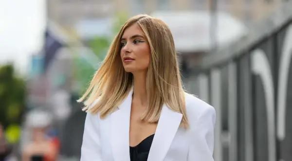 Frau mit blonden, leicht krisseligen Haaren nach Blondierung, trägt weißen Blazer und schwarzen BH.