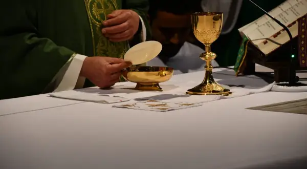 Kapłan trzyma hostię nad złotą pateną. Obok kielich i księga liturgiczna. To obrazuje przyczyny zmian w tekstach modlitw w nowym mszale rzymskim.
