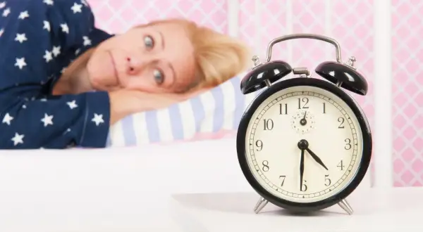 Mujer en pijama mira un despertador, su expresión refleja la **intención paradójica** de querer dormir pero no poder.