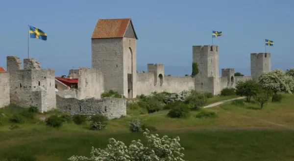 Visby muren reser sig stolt mot en blå himmel, med svenska flaggor som vajar från tornen. En historisk stadsvall som vittnar om svunna tider.