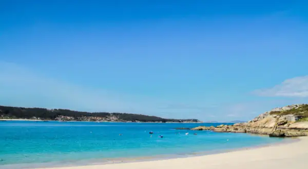 Arena blanca y aguas turquesas en la playa de Menduiña, con barcas a lo lejos y una costa rocosa.