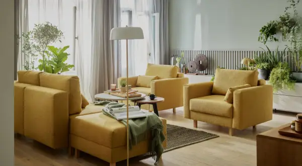 Przytulny salon bez telewizora. Żółte meble wypoczynkowe, stoliki kawowe, lampa podłogowa i mnóstwo zielonych roślin tworzą relaksującą atmosferę.