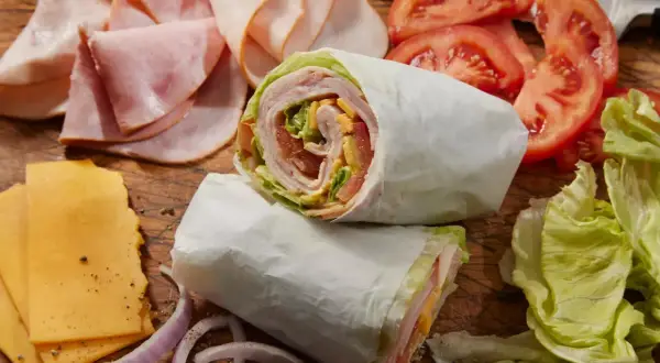 Dwa zawinięte w papier wrapy z szynką, serem, pomidorami i sałatą. Obok plastry szynki, sera, pomidorów i cebuli. Idealne na szybki posiłek, choć warto sprawdzić wrap mcdonald kcal.