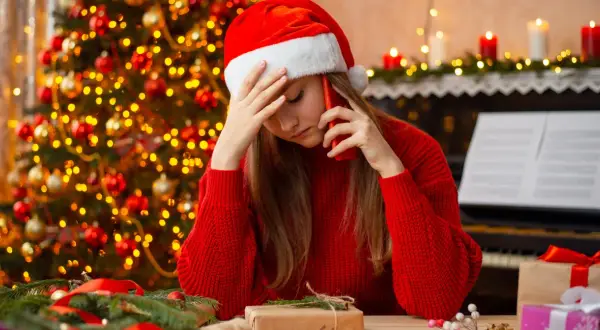 Smutna dziewczyna w czapce Mikołaja rozmawia przez telefon w Wigilię. W tle choinka i świece.