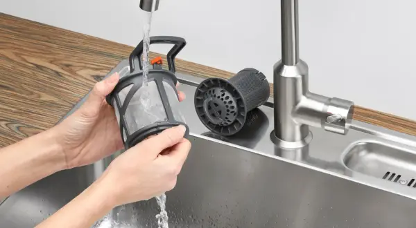 Czyszczenie zmywarki Electrolux. Ręce płuczą filtr pod bieżącą wodą w zlewie. Obok leży drugi, czysty filtr.