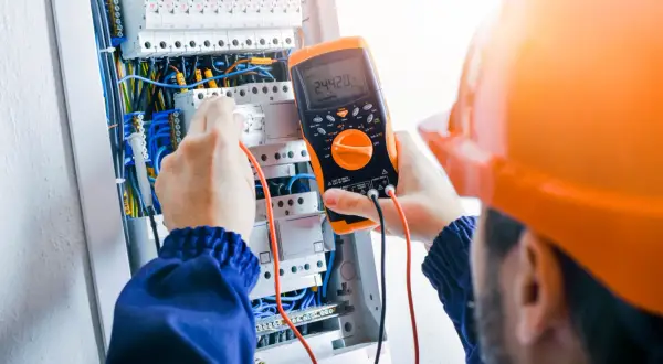 Elektryk w pomarańczowym kasku sprawdza napięcie multimetrem w skrzynce z przewodami.