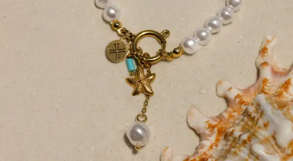 Un delicado llavero de oro con charms de estrella de mar y perla, sobre arena y junto a una concha marina.