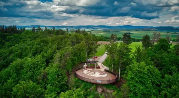 Widok na taras widokowy w otoczeniu lasu, z panoramą pól i gór w tle. Jakby to było jezioro wśród gór stołowych.