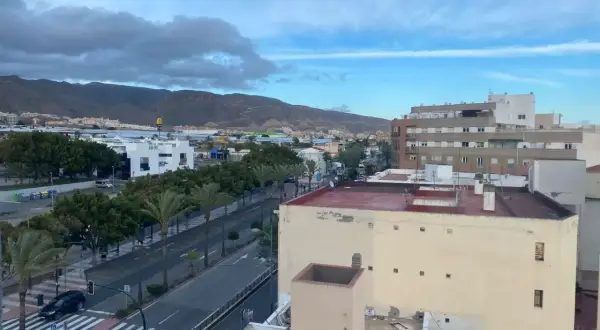 Vista aérea de una ciudad con edificios, palmeras y montañas al fondo. Las hortichuelas se ven en la distancia, bajo un cielo nublado.