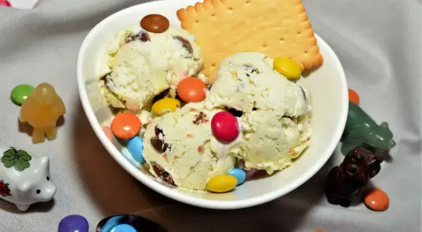 Schüssel mit cremigem Eis, garniert mit bunten Smarties und einem Keks. Kleine Figuren und weitere Smarties schmücken den Tisch.