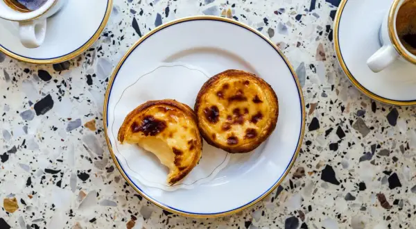 Dwa pyszne pastel de nata na talerzyku, obok filiżanki kawy. Idealny deser na każdą porę dnia.