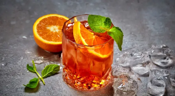 Un cocktail aranciato con fette d'arancia e menta, circondato da cubetti di ghiaccio per cocktail che non si scioglie, su uno sfondo grigio.