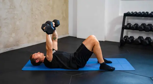 Uomo esegue esercizi pettorali con manubri su tappetino blu. Allenamento in palestra con rack di pesi sullo sfondo.