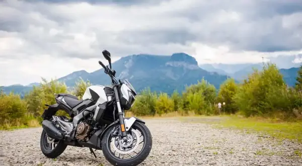 Biało-czarny motocykl Bajaj 400 na kamienistym podjeździe, w tle góry i chmury.