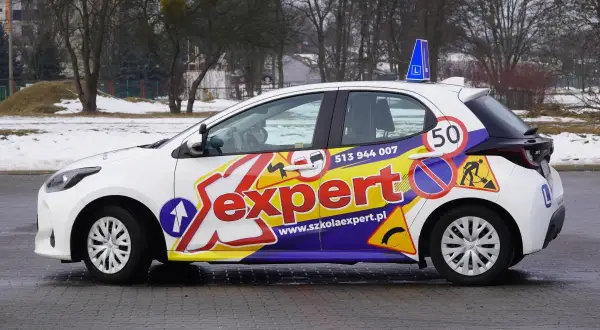 Biały samochód szkoleniowy z napisem "Sexpert" i znakami drogowymi, gotowy do prowadzenia pojazdu.