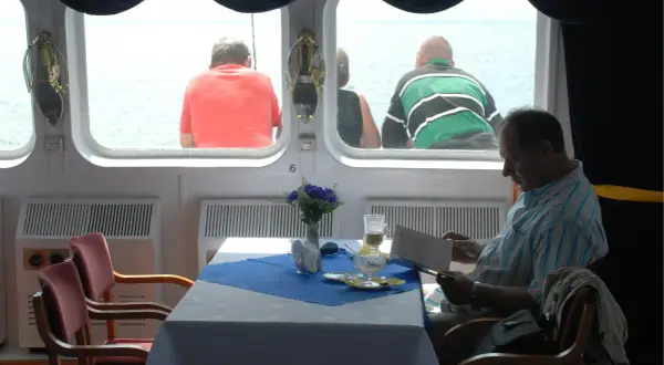Rejs PKP Gdynia - Hel. Cena biletu? Mężczyzna czyta w restauracji na statku, podczas gdy inni pasażerowie patrzą na morze.