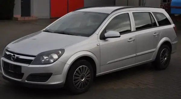 Srebrny Opel Astra H 1.6 benzyna, kombi, zaparkowany na kostce brukowej przed budynkiem z czerwonymi drzwiami.