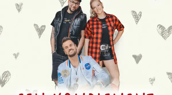Ein Mann und zwei Frauen, eine girls group, posieren lächelnd vor einem Hintergrund mit Herzen.