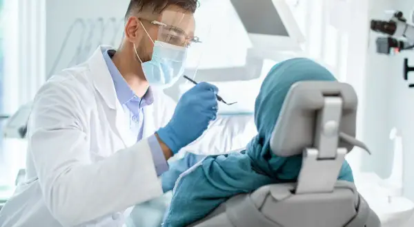 Lekarz stomatolog w masce i fartuchu bada pacjentkę w chuście. Nawet przy dentofobii, wizyta u dentysty jest ważna.