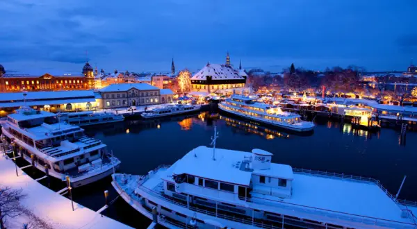 Schneebedeckte Schiffe im Hafen von Konstanz, beleuchtet vom Glanz des Weihnachtsmarktes.