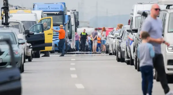 Stau auf der Autobahn. Ein Mann in orangefarbener Jacke und Hut steht neben einem gelb-schwarzen Transporter, dessen Tür offen ist. Viele Autos und LKW stehen still, als ob sie trotz Fahrverbot unterwegs wären.