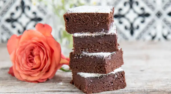 Schokoladige Brownies mit Datteln, bestäubt mit Puderzucker, neben einer zarten orangefarbenen Rose auf Holztisch.
